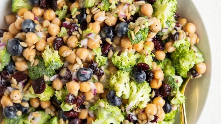 Big white bowl of broccoli chickpea salad with a gold spoon and a bowl of sunflower seeds.