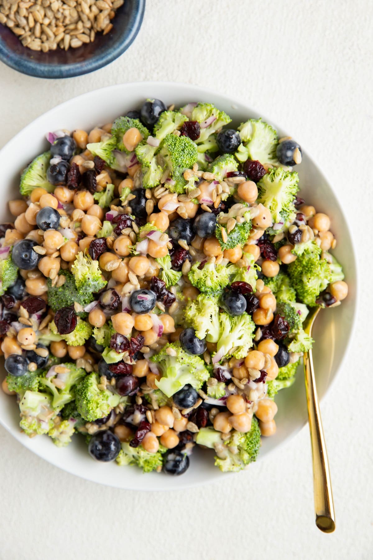 Big white bowl of broccoli chickpea salad with a gold spoon and a bowl of sunflower seeds.