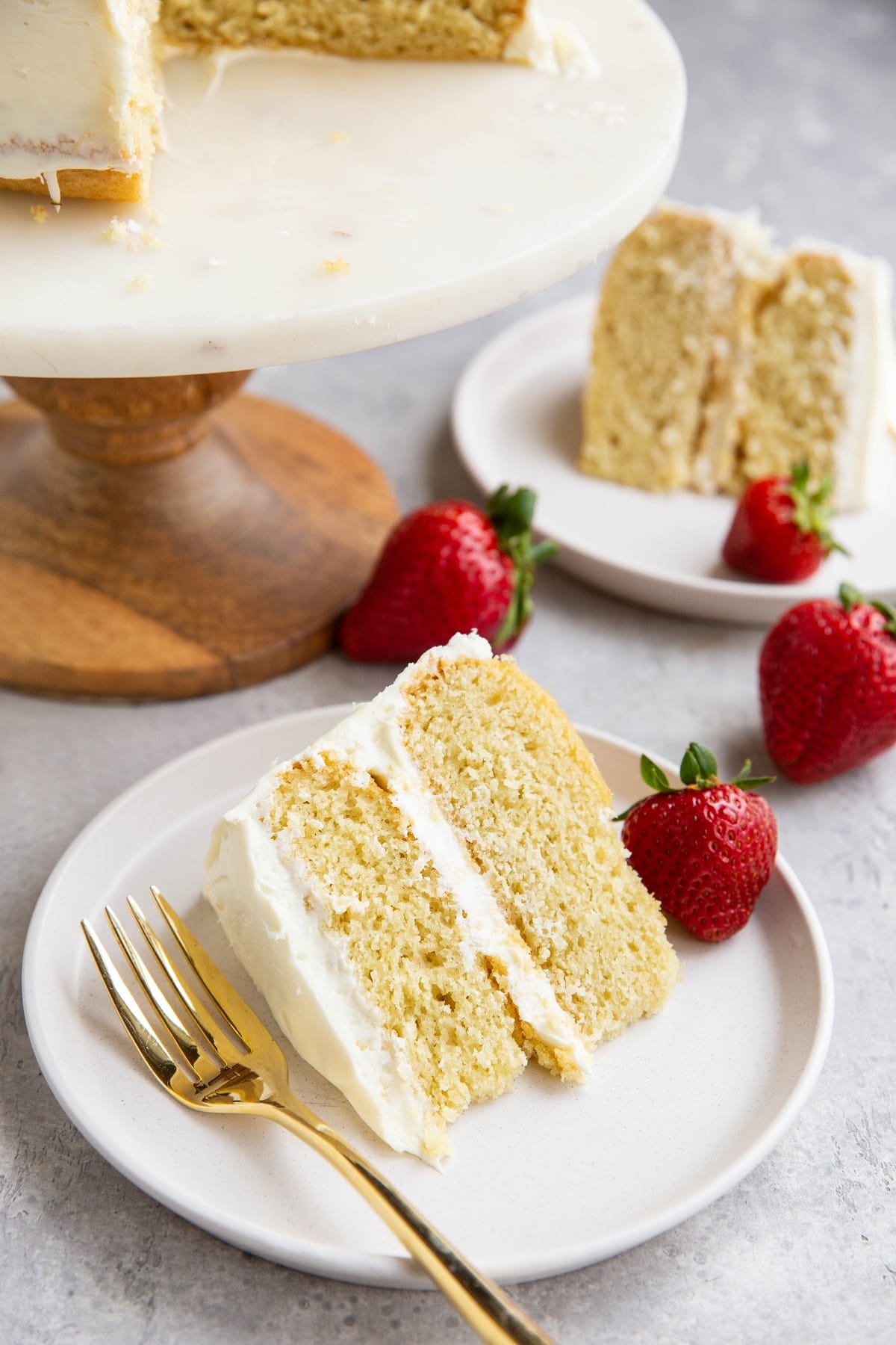 Two slices of vanilla cake with a cake stand with the rest of the cake in the background.