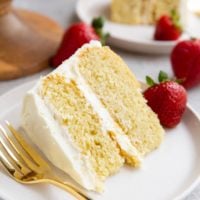 Two slices of vanilla cake on two white plates with fresh strawberries to the side.