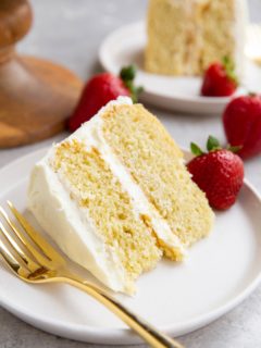 Two slices of vanilla cake on two white plates with fresh strawberries to the side.