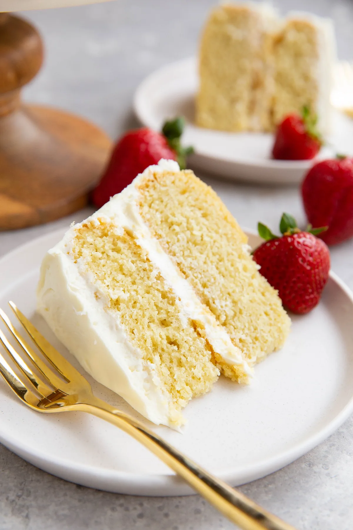 Two slices of vanilla cake on two white plates with fresh strawberries to the side.