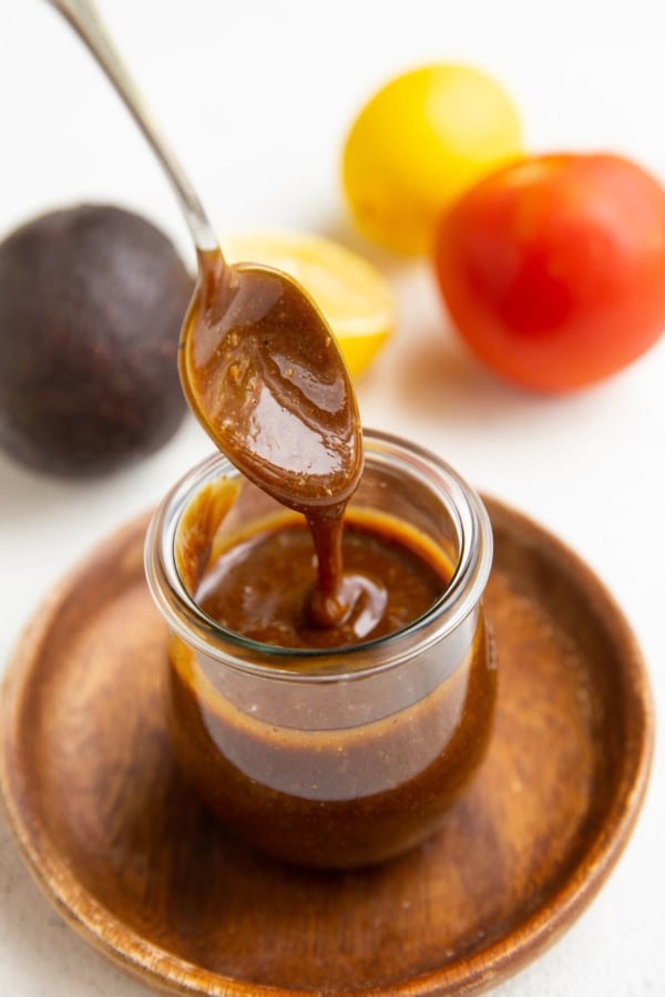 Spoon drizzling balsamic vinaigrette in a jar with fresh produce in the background.