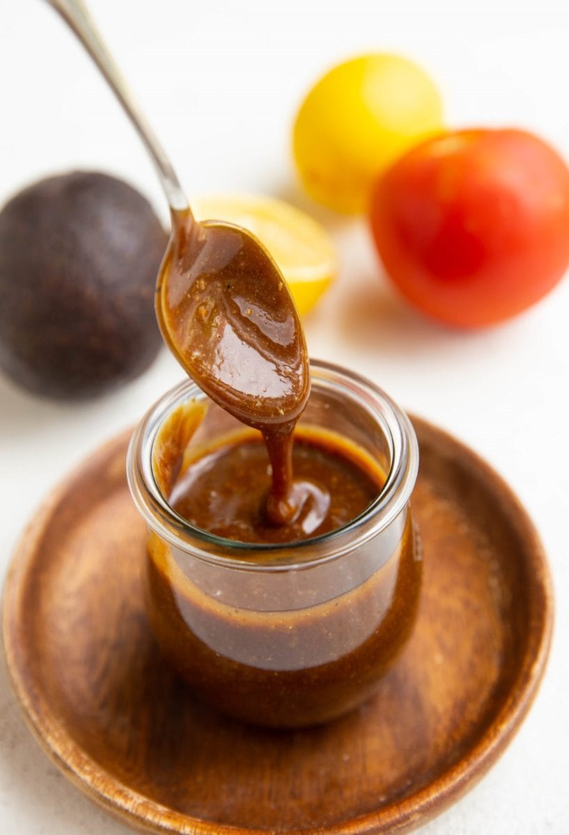 Spoon drizzling balsamic vinaigrette in a jar with fresh produce in the background.