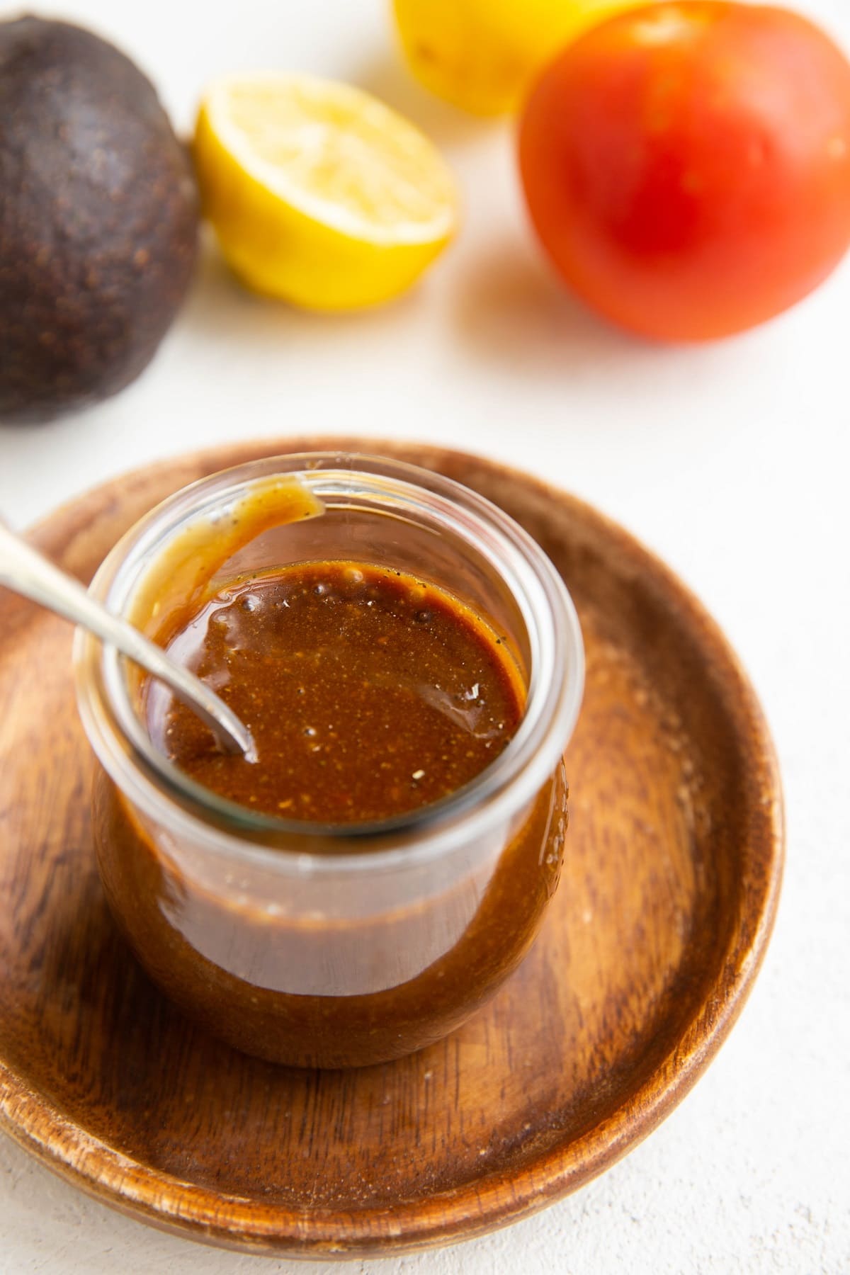 Jar of balsamic vinaigrette with a tomato, lemon, and avocado in the background.