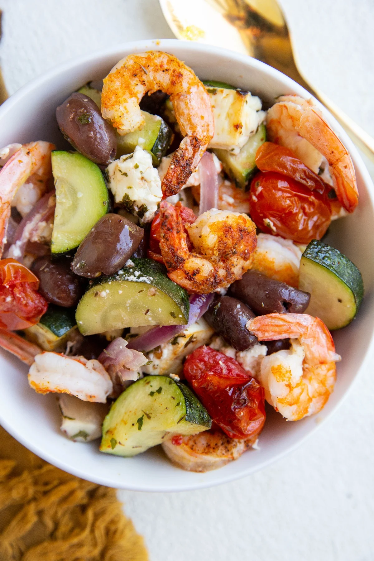 White bowl of shrimp and vegetables with feta and olives. A golden spoon to the side, ready to eat.