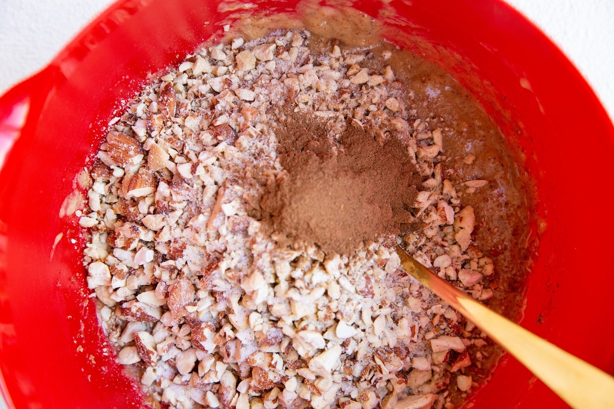 Almond butter, mashed banana at the bottom of a mixing bowl with chopped raw nuts, cinnamon and salt on top, ready to be mixed in.