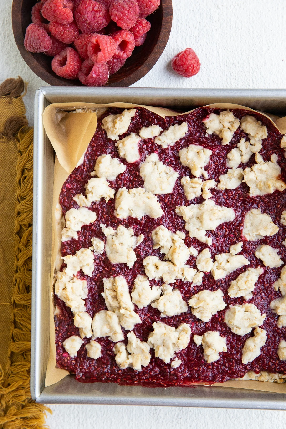 Square baking pan with raspberry crumb bars inside. A bowl of fresh raspberries and a gold napkin to the side of the baking dish.
