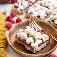 Wooden plate with a slice of raspberry crumb bar and a gold fork. More crumb bars in the background.