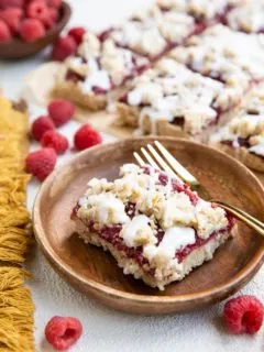 Wooden plate with a slice of raspberry crumb bar and a gold fork. More crumb bars in the background.