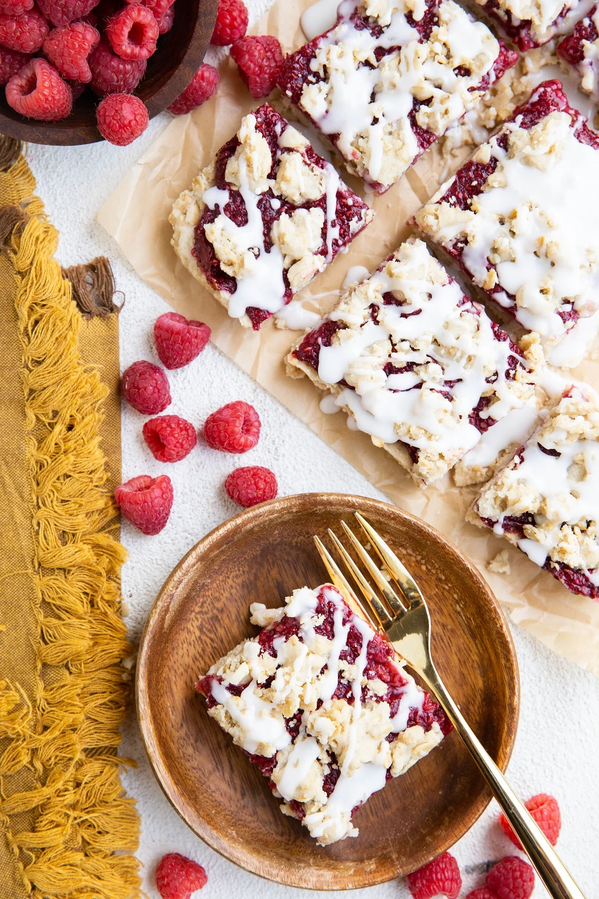 Parchment paper with slices of raspberry crumb bars and a wooden plate with a crumb bar. Fresh raspberries around the sides and a golden napkin.