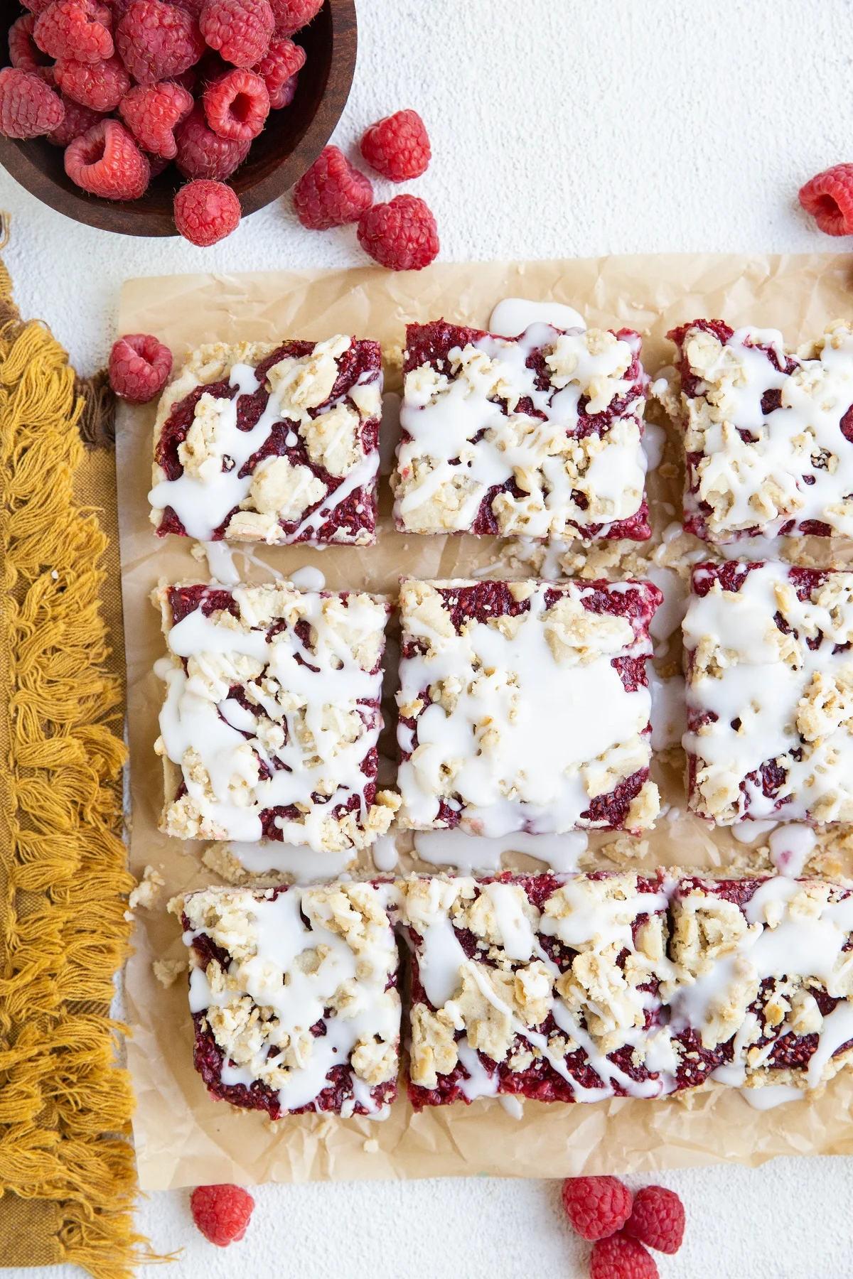 Raspberry crumb bars cut into squares on a sheet of parchment paper with fresh raspberries around.