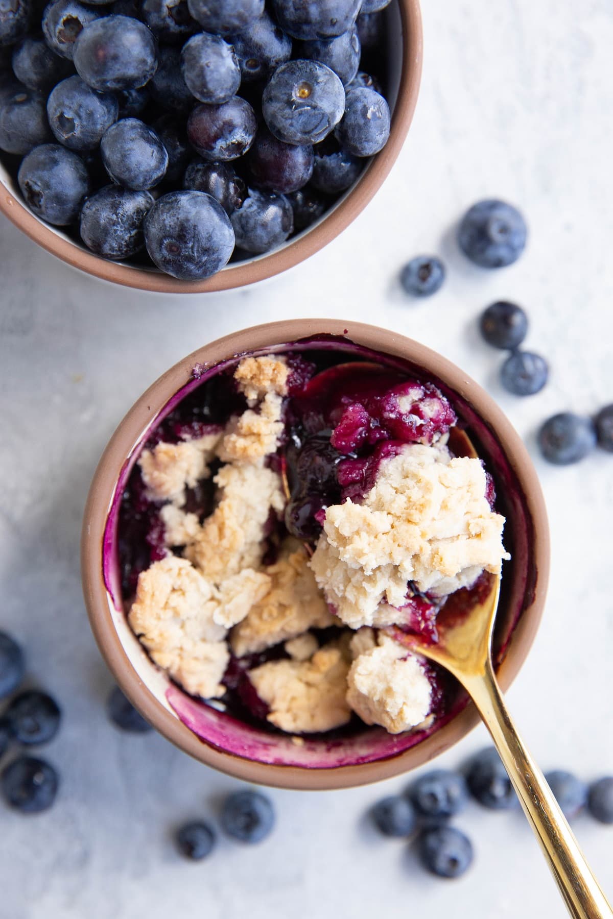 Ramekin of blueberry cobbler with a ramekin of fresh berries. Golden spoon with a bite, ready to eat.