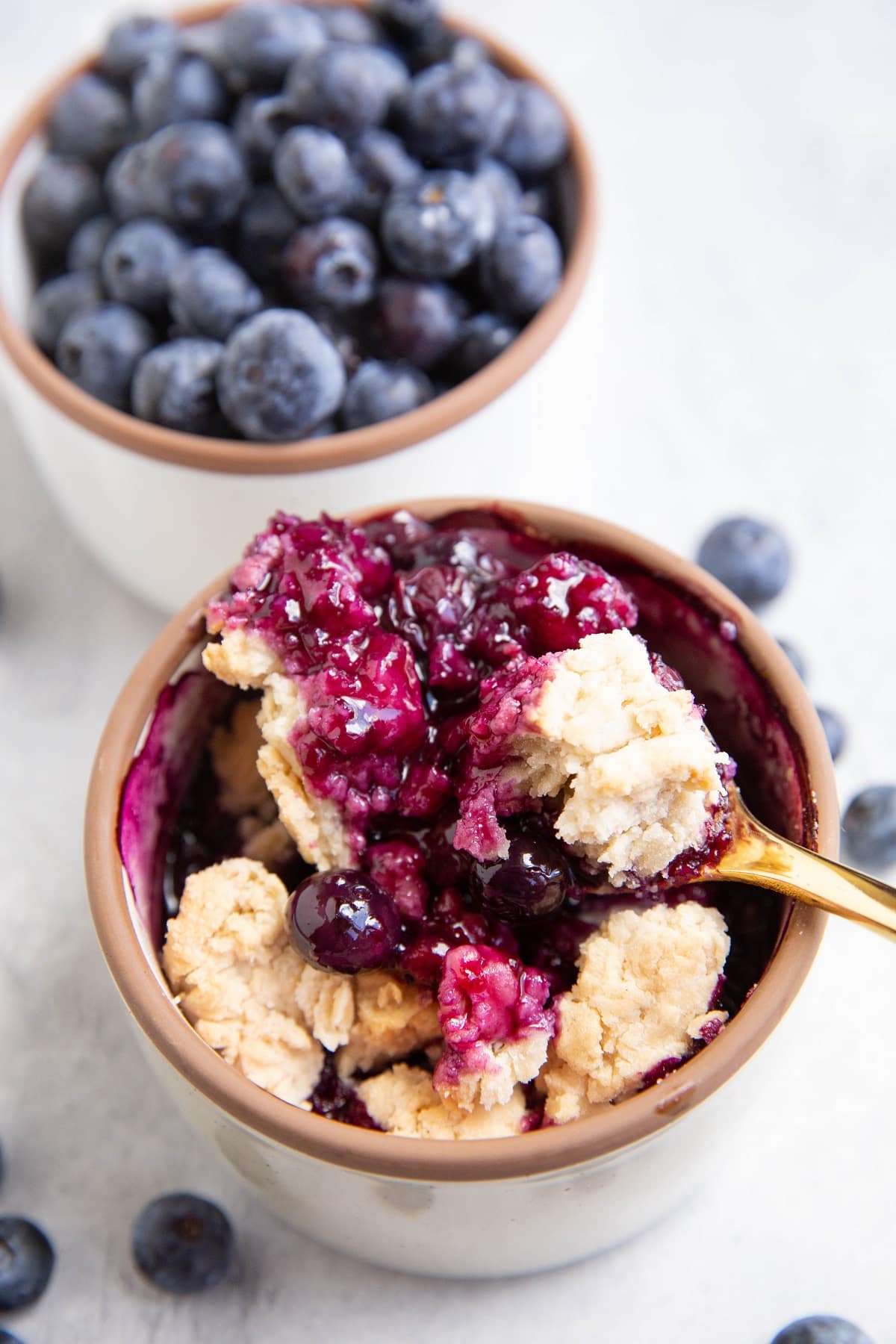 White ramekin with blueberry cobbler and a bowl of fresh blueberries in the background.