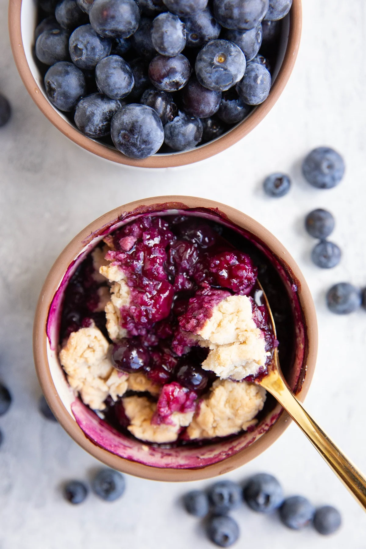 Two ramekins of blueberries and blueberry cobbler with fresh blueberries all around.