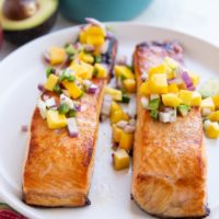 Two cooked salmon filets on a white plate with mango salsa on top.