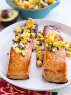 Two cooked salmon filets on a white plate with mango salsa on top.