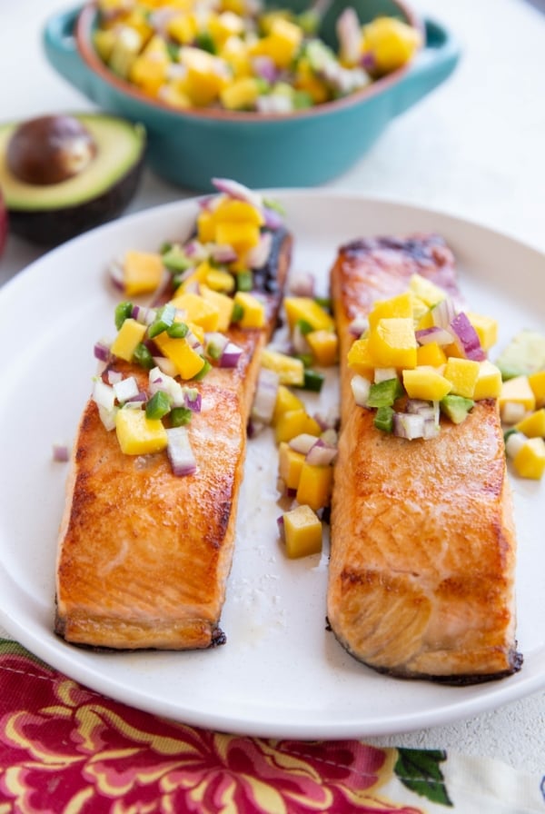 Two cooked salmon filets on a white plate with mango salsa on top.