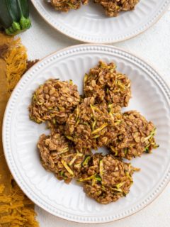 Two white plates of zucchini oatmeal cookies with a golden napkin and a fresh zucchini to the side.