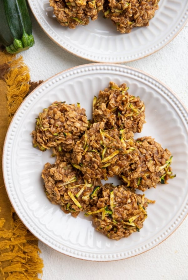 Two white plates of zucchini oatmeal cookies with a golden napkin and a fresh zucchini to the side.