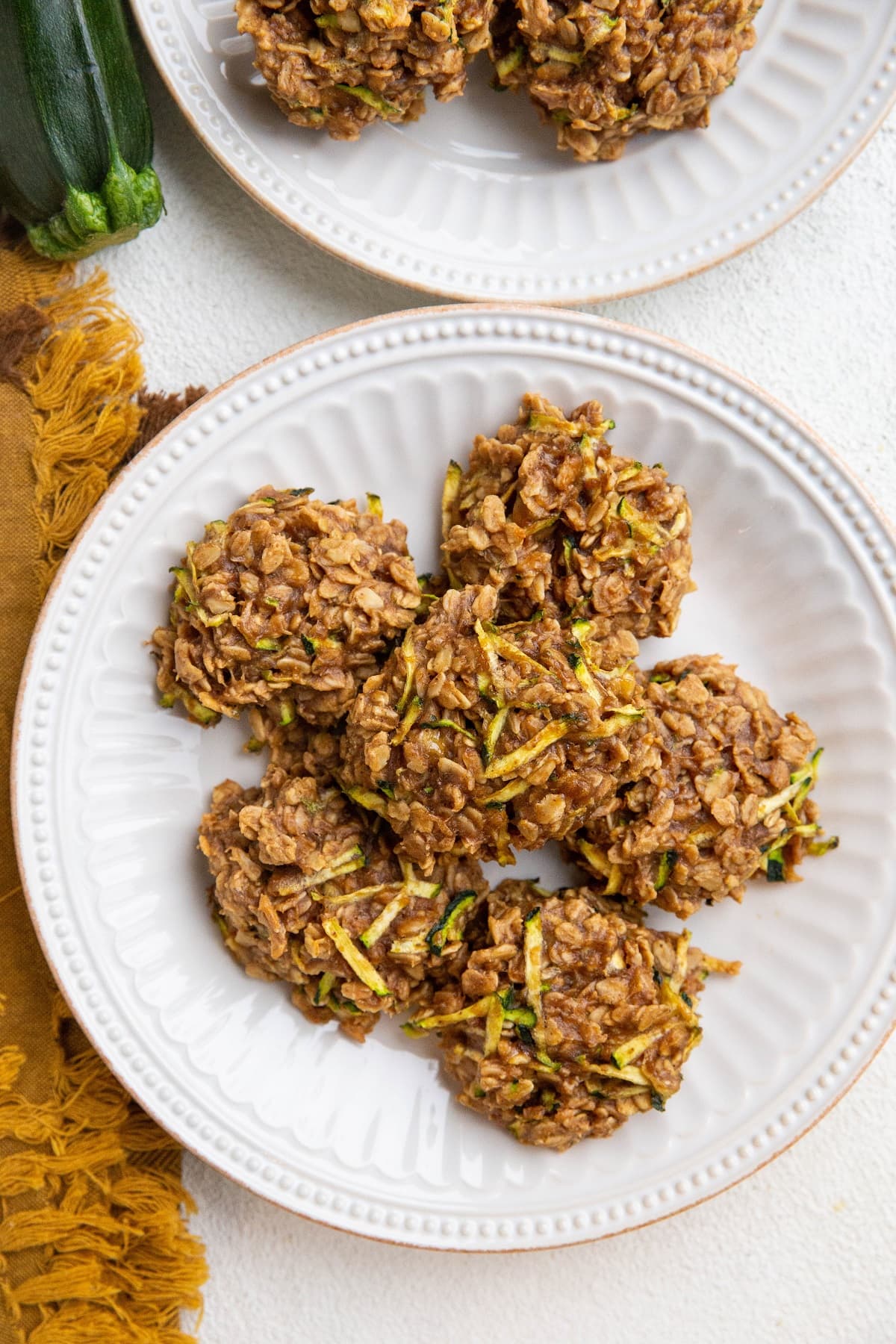 Two white plates of zucchini oatmeal cookies with a golden napkin and a fresh zucchini to the side.