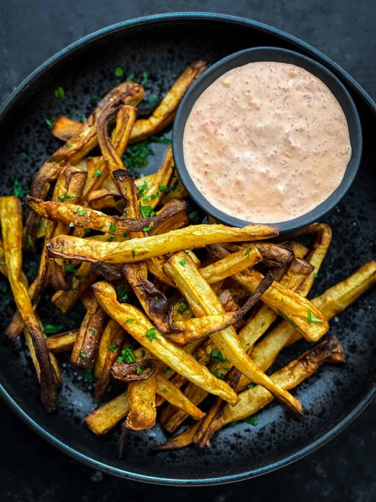 Parsnip fries on a black plate with a dipping sauce to the side