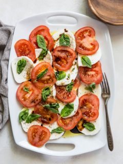 White serving platter of caprese salad with a wooden plate and a serving fork.