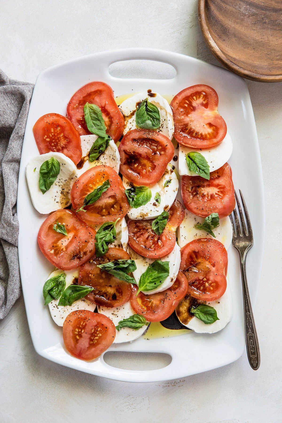 White serving platter of caprese salad with a wooden plate and a serving fork.