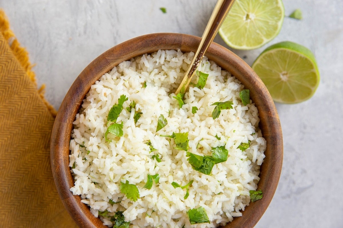 Wooden bowl of cilantro lime rice with a golden serving spoon and fresh limes and a golden napkin to the side.