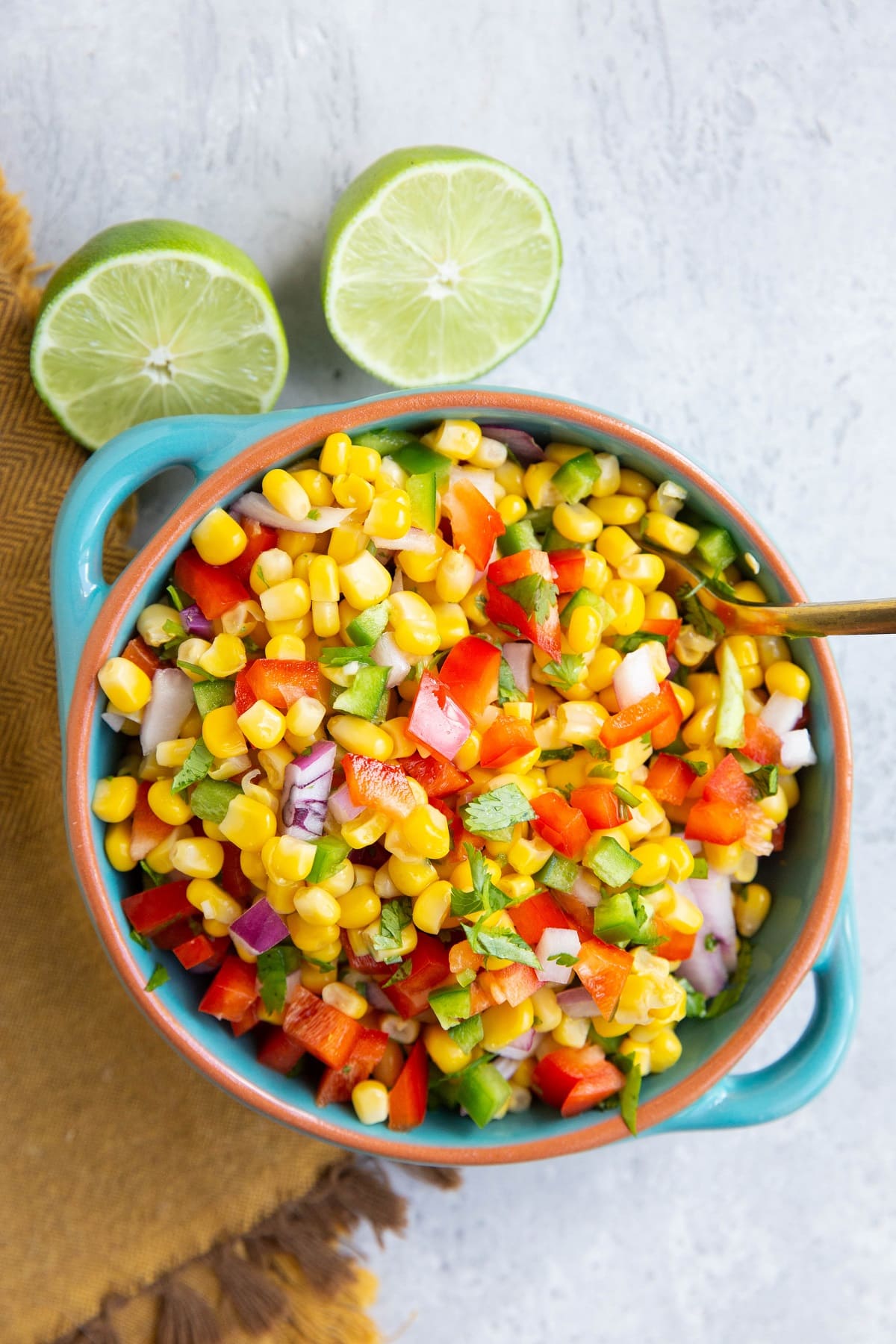 Bowl of corn salsa with a gold serving spoon, golden napkin and lime to the side.