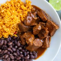 White bowl of Chile Colorado, rice, beans, and a fresh lime to the side.