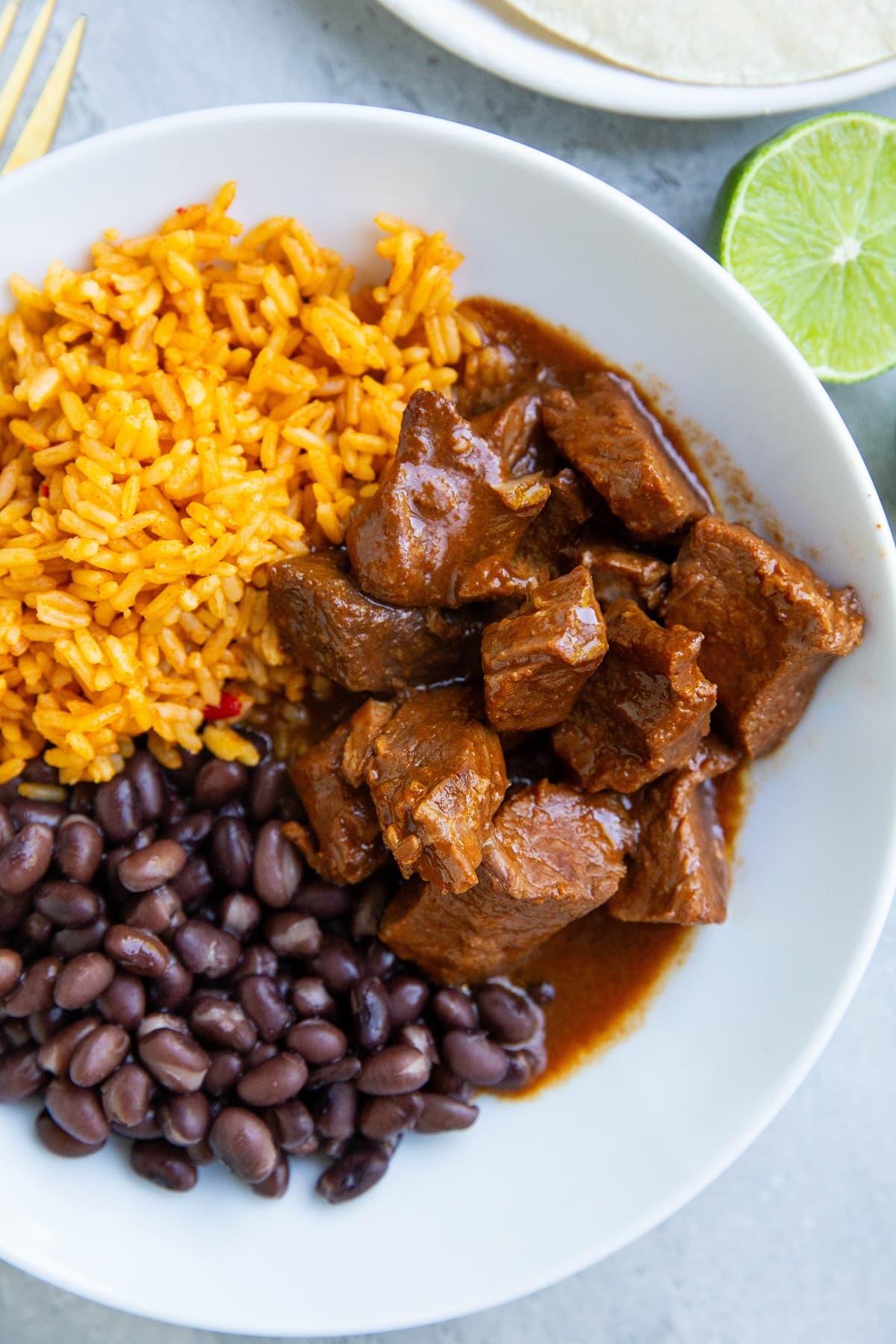 White bowl of Chile Colorado, rice, beans, and a fresh lime to the side.