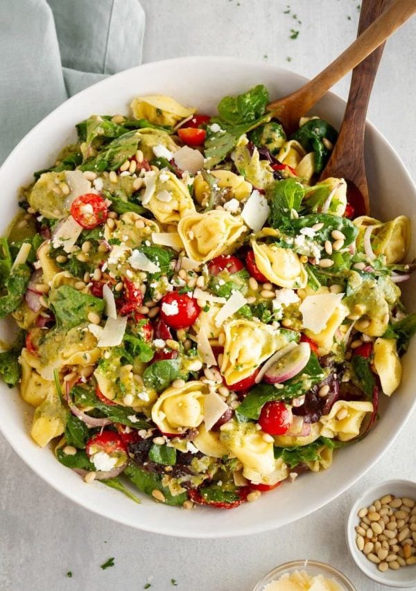 Large bowl of tortellini salad with wooden spoons, pine nuts and parmesan cheese to the side.
