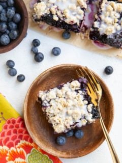 Blueberry crumb bar on a wooden plate with a bowl of blueberries and a napkin and fork around.