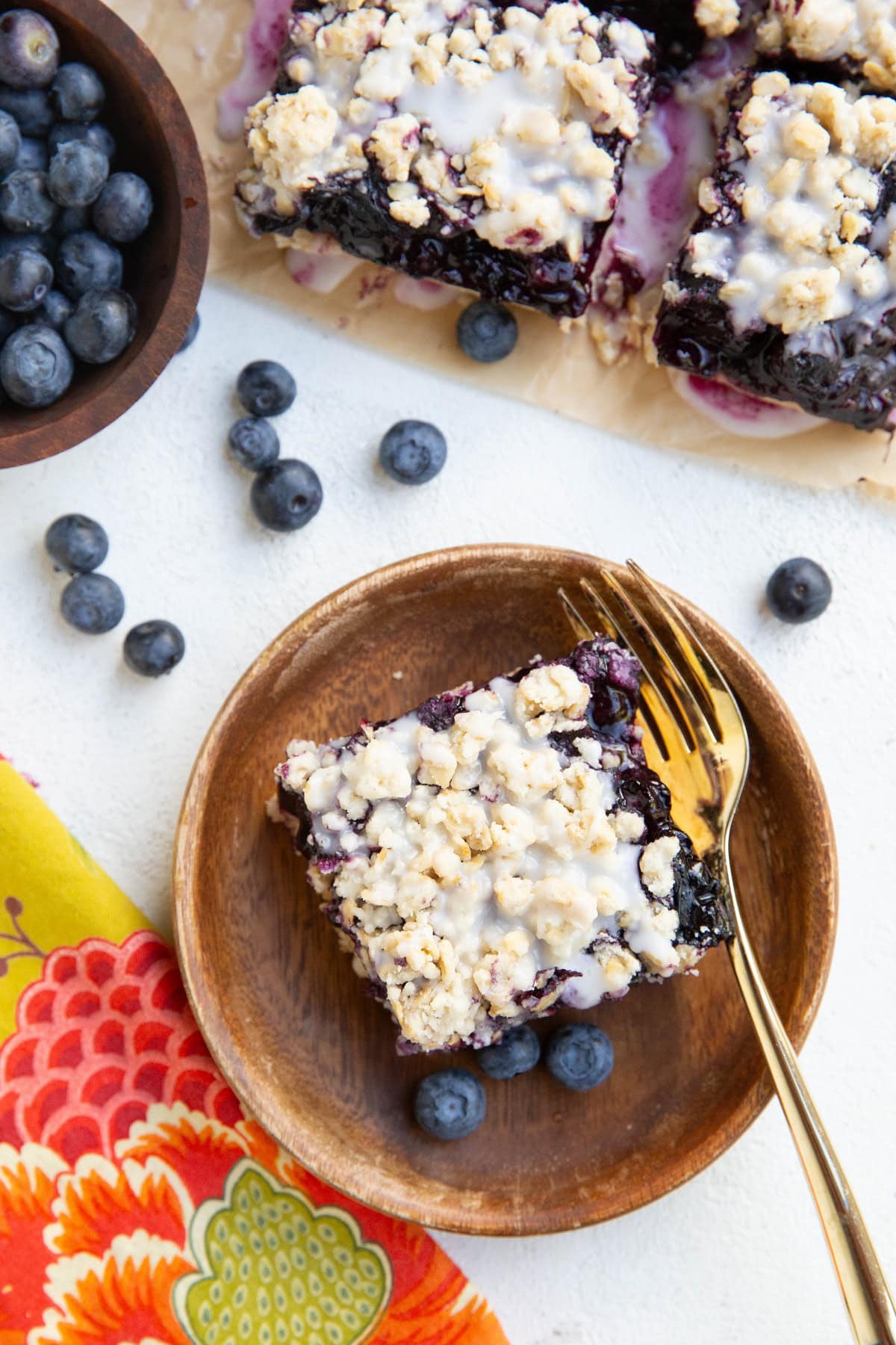 Blueberry crumb bar on a wooden plate with a bowl of blueberries and a napkin and fork around.