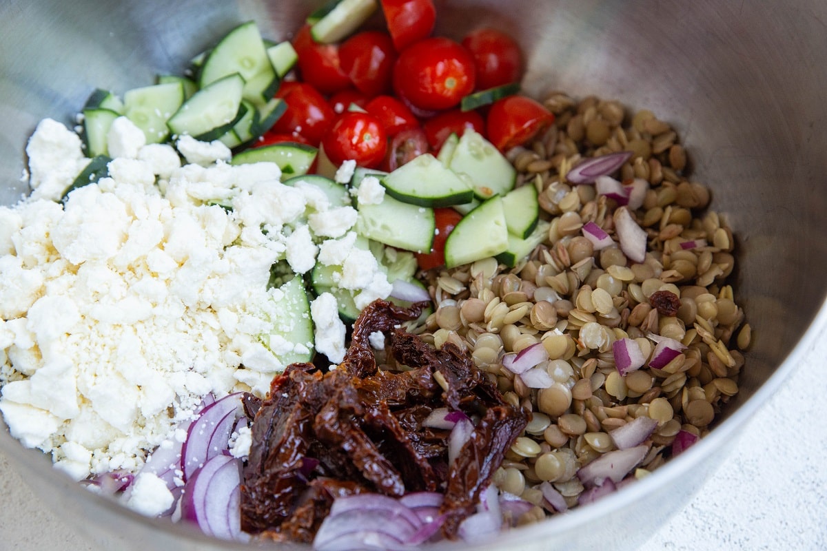 lentils, tomatoes, cucumber, raw onions, sun-dried tomatoes, and feta cheese in a mixing bowl.
