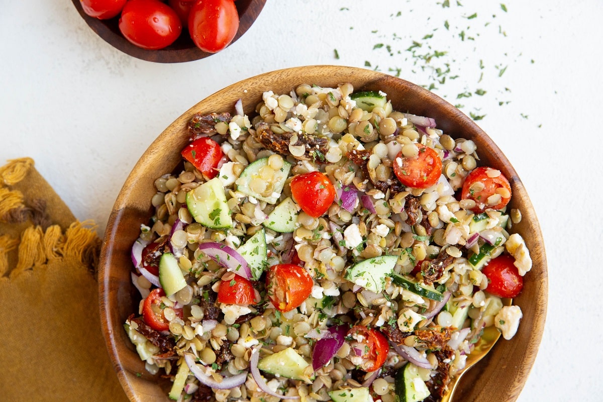 Wooden bowl full of Greek lentil salad with cherry tomatoes to the side and a golden napkin and dried parsley.