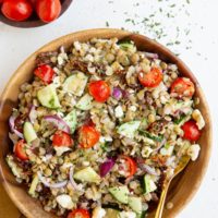 Wooden bowl of lentil salad with a golden spoon and fresh tomatoes to the side.