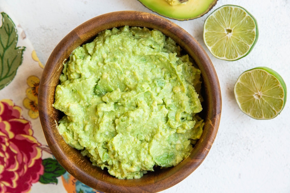 Wooden bowl of guacamole with fresh limes and avocado to the side.