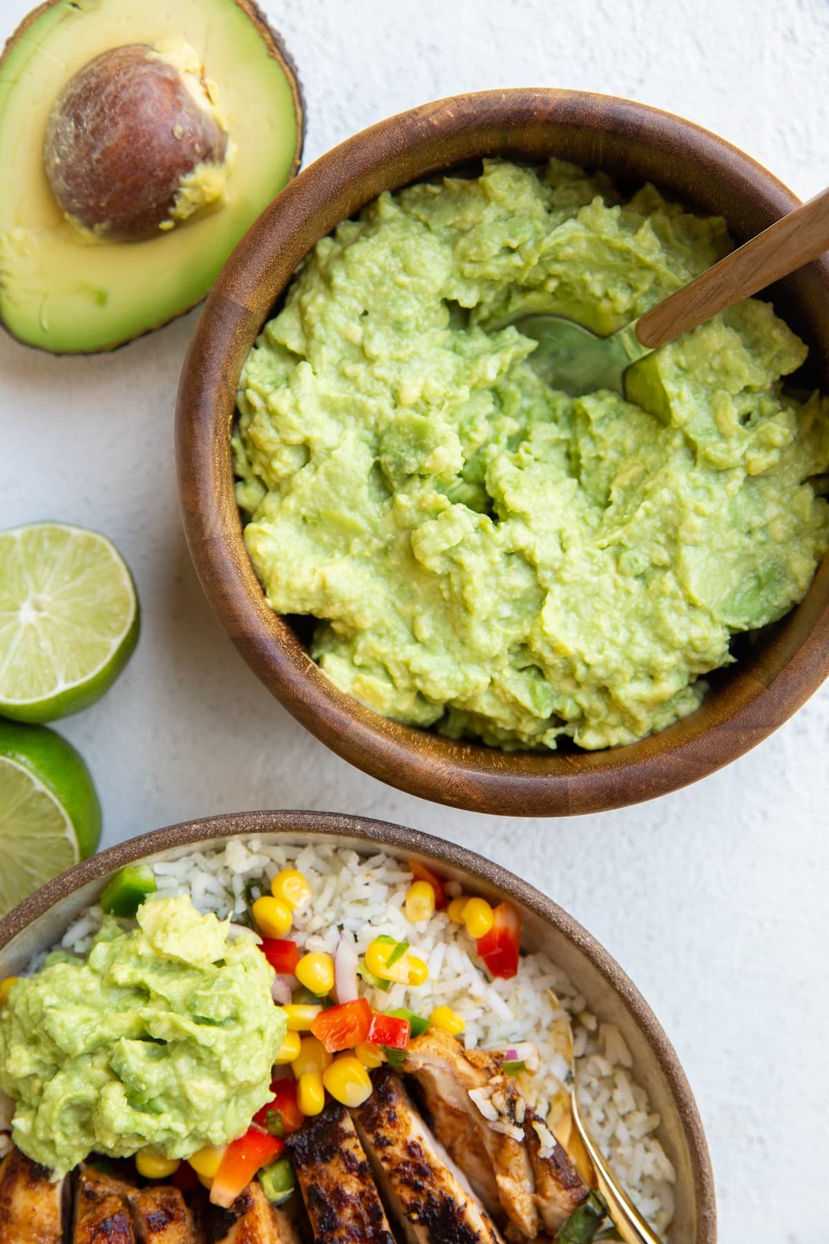 Wooden bowl of guacamole with a chicken burrito bowl to the side with guacamole on top.