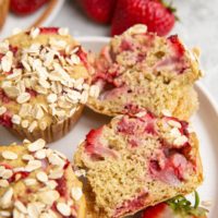 white plate of strawberry oatmeal muffins with one sliced in half and fresh strawberries in the background.