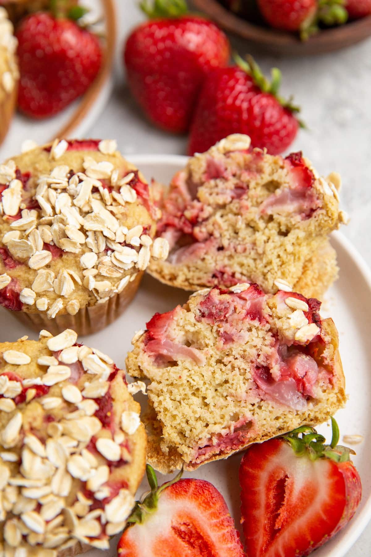 white plate of strawberry oatmeal muffins with one sliced in half and fresh strawberries in the background.