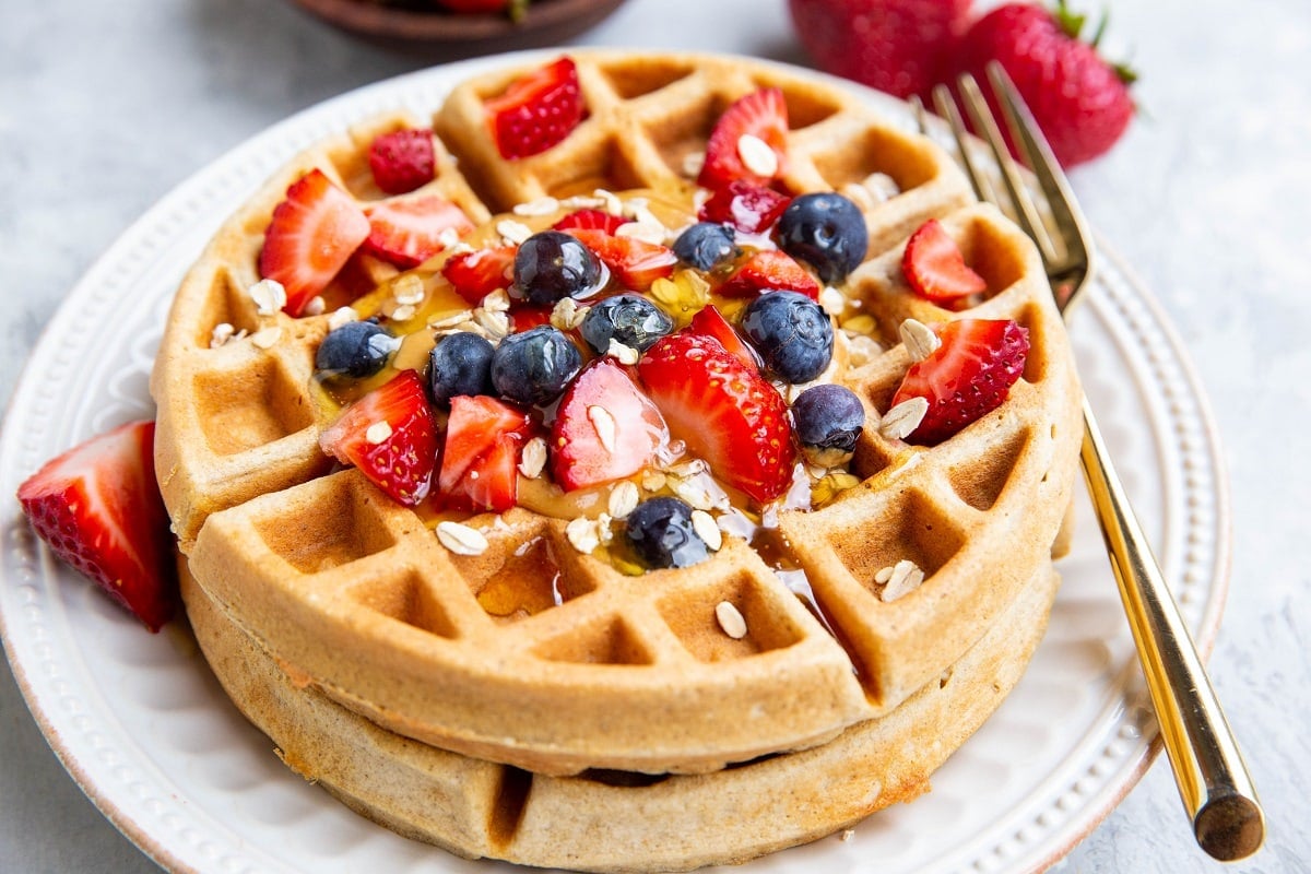 Stack of two Belgian waffles on a white plate with berries and honey on top with a gold fork.