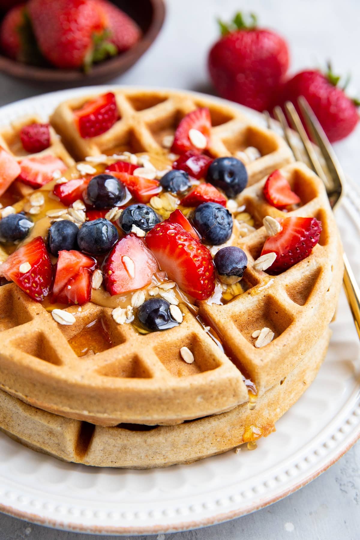 Stack of protein waffles on a white plate with fresh fruit, syrup, and nut butter.
