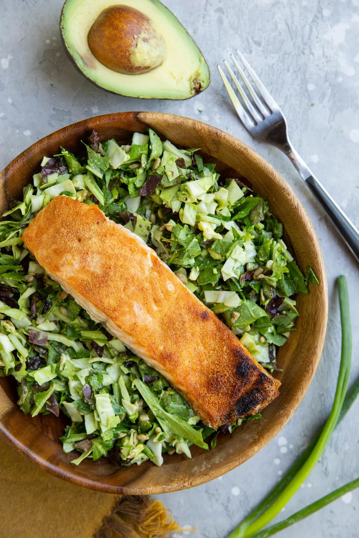 Wooden salad bowl with salad and salmon with an avocado and green onion to the side.