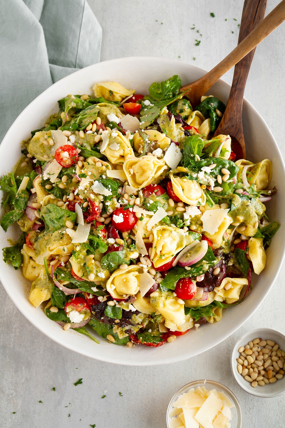 Large bowl of tortellini salad with wooden spoons, pine nuts and parmesan cheese to the side.