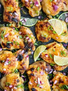 Chicken thighs in a casserole dish, sprinkled with green onions, red onions and lime slices.