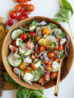 Big wood serving bowl with cucumber tomato salad inside with fresh tomatoes and basil to the side.