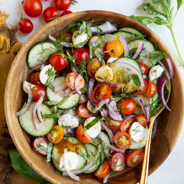 Big wood serving bowl with cucumber tomato salad inside with fresh tomatoes and basil to the side.