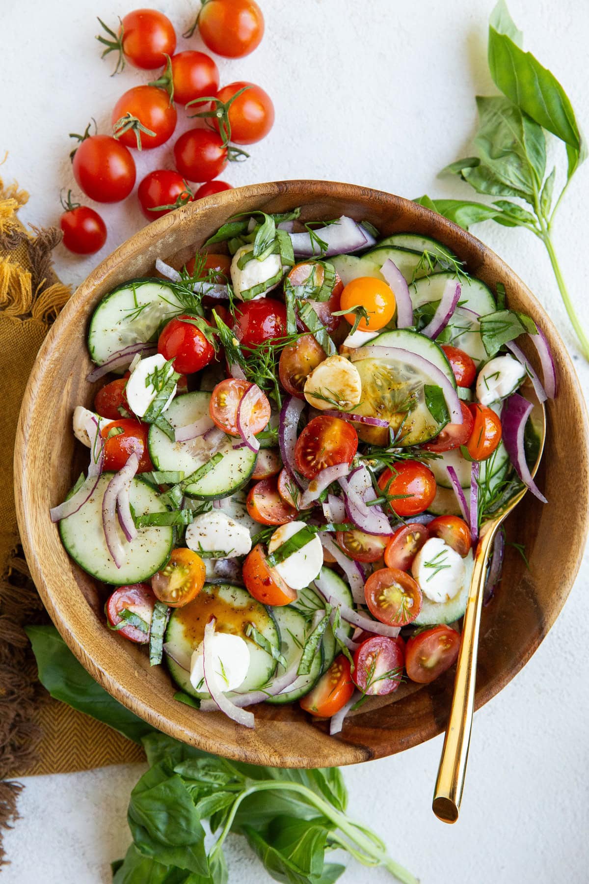 Big wood serving bowl with cucumber tomato salad inside with fresh tomatoes and basil to the side.